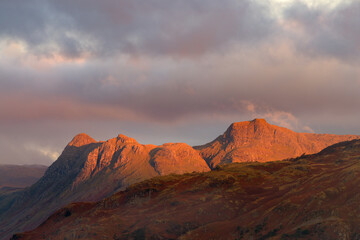 Naklejka premium Beautiful red sunlight from the rising sun illuminating the Langdale Pikes mountain range in the English Lake District with dark clouds in sky.
