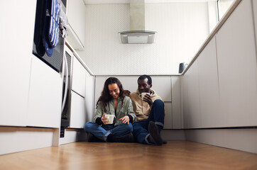 Young Couple Celebrate Moving Into New Home Sitting On Floor With Hot Drink In Kitchen