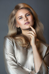 A portrait of a sexy young white female with long blond curly hair, beautiful makeup posing by herself in a studio with a grey background wearing a beautiful silver dress.