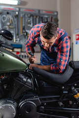 Plakat young mechanic holding socket wrench while examining motorcycle in workshop