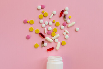 Assorted medical pills and capsules scattered from a white plastic bottle on a pink background.