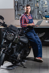 Fototapeta na wymiar mechanic in overalls looking at camera while standing near motorbike with laptop