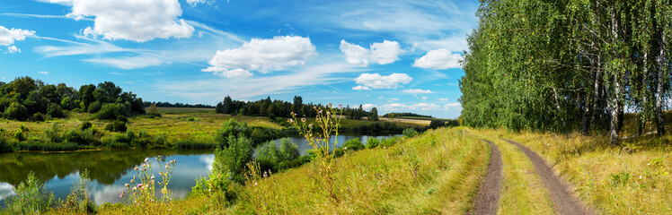 Sunny summer rural landscape with calm river, birch trees near country road and farm fields