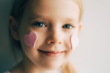 Close-up portrait of a girl wirh hearts on her face. Pink hearts on  cheeks. Valentines concept. Portrait of a blonde child.  - 403066814