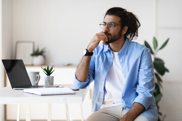 Thoughtful young arab male freelancer sitting at desk at home, looking aside - Powered by Adobe