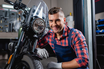 cheerful mechanic in overalls smiling at camera near motorbike in workshop