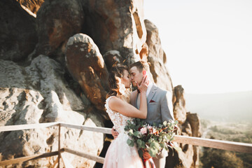 Young newly wed couple, bride and groom kissing, hugging on perfect view of mountains, blue sky