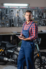 young technician in overalls looking at camera while standing near motorcycle with digital tablet, blurred background