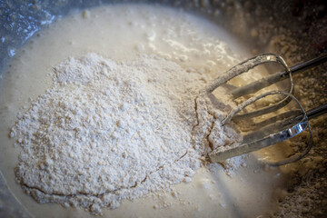 Electric mixer beaters resting in bowl of baking batter with flour on top that needs to be beaten in - selective focus and close-up