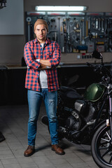 Fototapeta na wymiar full length view of mechanic in plaid shirt and jeans standing with crossed arms near motorcycle in workshop