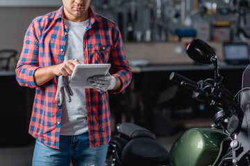 partial view of mechanic in checkered shirt using digital tablet near motorcycle in workshop