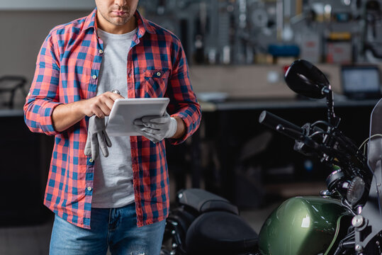 partial view of mechanic in checkered shirt using digital tablet near motorcycle in workshop - Powered by Adobe