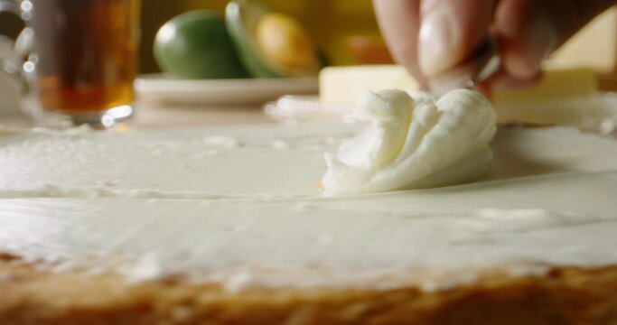Close up shot of a knife spreading cream cheese on a piece of bread, tasty food being prepared for breakfast or lunch with kitchen on background 4k footage