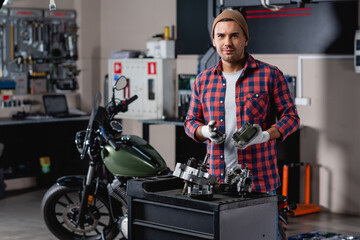 Fototapeta na wymiar smiling mechanic in plaid shirt and gloves holding gearwheels near disassembled motorbike transmission