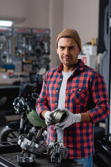 young mechanic in checkered shirt and beanie looking at camera near disassembled motorcycle gearbox