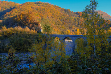 old stone bridge over the mountain river across autumn forest, hills, and sunset sky. Natural background. Travel, movement, architecture concept