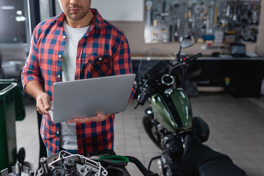 cropped view of mechanic using laptop near spare parts and motorcycle on blurred background