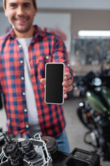 smiling mechanic showing smartphone with blank screen near motorcycle spare parts, blurred background