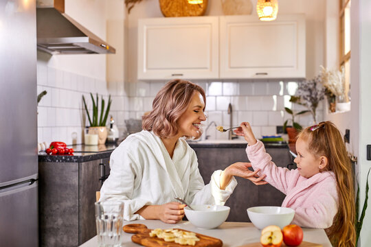 mother with kid girl eating tasty porridge for breakfast, daughter is treating mother by spoon, in light kitchen at home