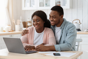 Smiling black couple with laptop on kitchen calculating bills, managing finances together