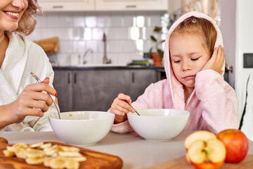 child girl doesn't want eat breakfast, sit behind table want to sleep, in light modern kitchen in the morning