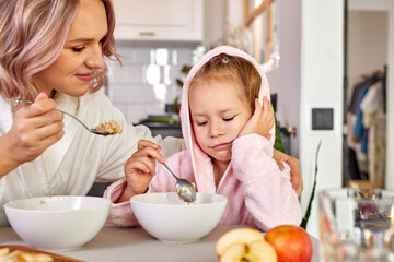 Obraz na płótnie Canvas mother persuades her daughter to eat at least a spoonful of porridge, little girl is dissatisfied, sad, sits wants to sleep