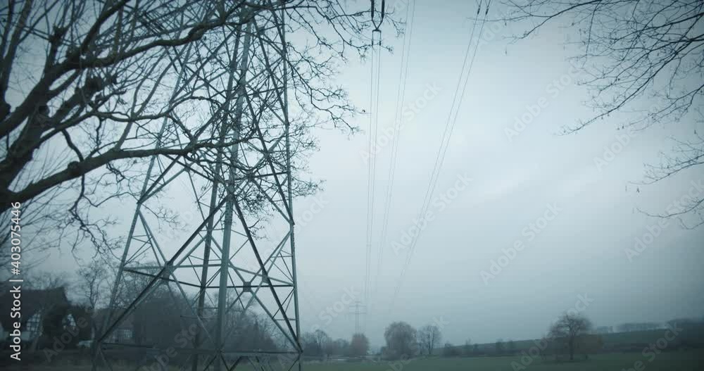 Poster electricity pylons in a field with bare trees in front of farmhouses in cloudy weather with grey sky