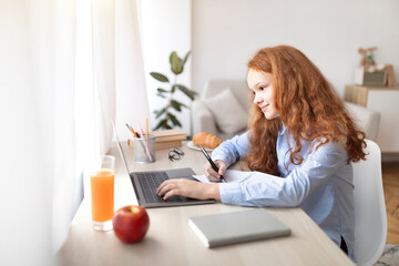Girl sitting at table, using laptop, writing in her notebook