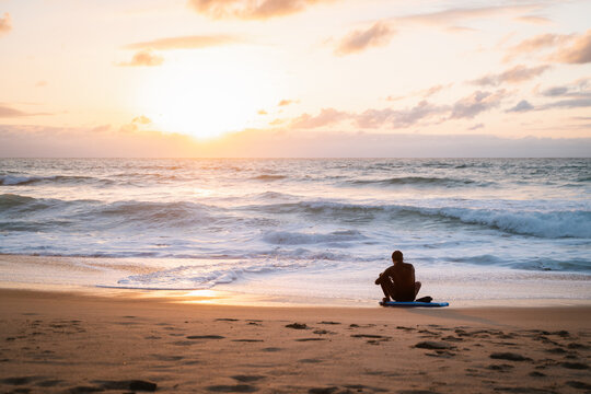 Young man sitting on his body board on the shore of a lonely beach during sunset adjusting his wet suit - Powered by Adobe