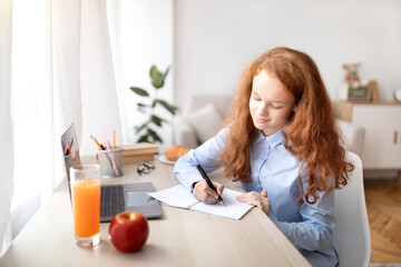 Girl sitting at table, using laptop, writing in her notebook