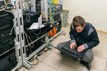 Young guy is sitting on the floor in the server room. A system administrator with a laptop works in...