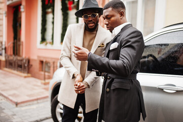Two fashion black men stand near business car and look at cell phone. Fashionable portrait of african american male models. Wear suit, coat and hat.