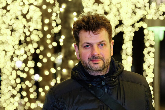 A man is posing for a chest-up portrait. It’s late at night and id dark outside. The Christmas Lights are in abundance in the background and a telephoto lens transformed them into a beautiful bokeh.