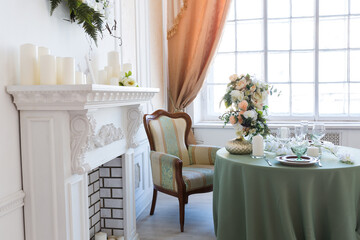 Luxury stylish bright light interior of sitting room. White walls decorated by ornament. Fireplace. Nobody inside room. Table setting by dishes, candles and flower bouquets.