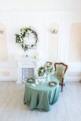 Luxury stylish bright light interior of sitting room. White walls decorated by ornament. Fireplace. Nobody inside room. Table setting by dishes, candles and flower bouquets.