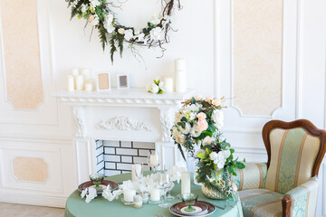 Luxury stylish bright light interior of sitting room. White walls decorated by ornament. Fireplace. Nobody inside room. Table setting by dishes, candles and flower bouquets.