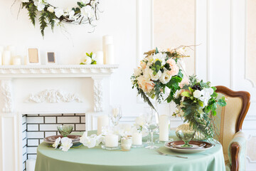 Luxury stylish bright light interior of sitting room. White walls decorated by ornament. Fireplace. Nobody inside room. Table setting by dishes, candles and flower bouquets.