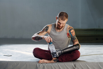 Cyborg man with bionic hand sitting on a mat, opening plastic bottle with water, taking a break. He...