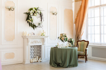 Luxury stylish bright light interior of sitting room. White walls decorated by ornament. Fireplace. Nobody inside room. Table setting by dishes, candles and flower bouquets.