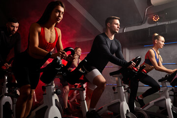 men and women biking in gym, exercising legs doing cardio workout cycling bikes, spinning in health club, wearing tracksuit sportive outfit