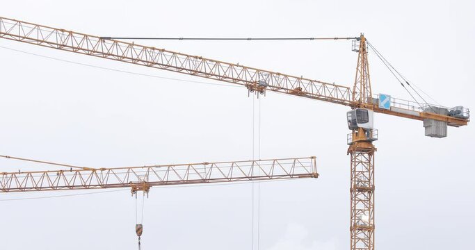 Two yellow tower crane jibs with working trolley on a construction site against white cloudy sky
