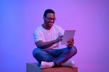 Young black freelancer guy sitting with digital tablet under luminous neon light - obrazy, fototapety, plakaty