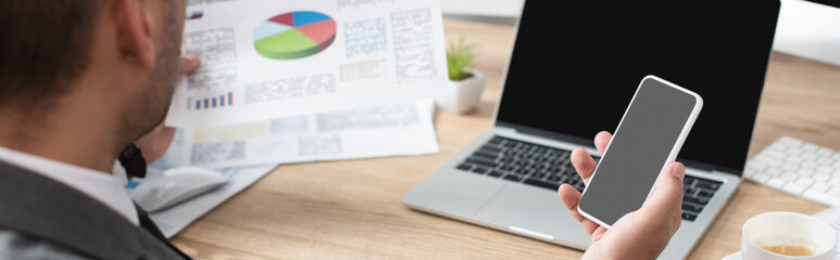 partial view of trader holding smartphone with blank screen and papers with charts on blurred foreground, banner