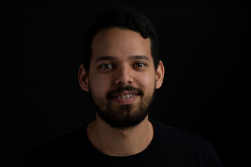 Obraz premium Young man frontal head shot with black background. He is smiling. He has a casual look with beard and t-shirt.