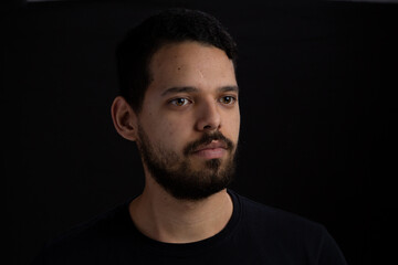 Young man head shot with black background. He is serious. He has a casual look with beard and t-shirt.
