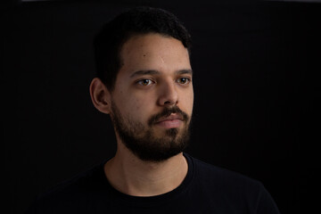 Obraz premium Young man head shot with black background. He is serious. He has a casual look with beard and t-shirt.