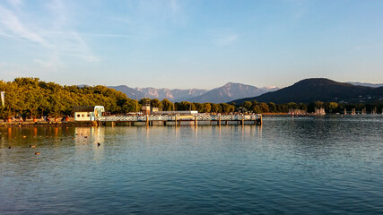 Fototapeta premium A panoramic capture of the pier on the Woerthersee lake in Austria. In the back there are high Alpine chains. The shore of the lake is overgrown with dense forest. Serenity and calmness. Golden hour.