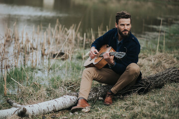 Naklejka na ściany i meble caucasian bearded man is playing guitar near the lake. handsome stylish hipster