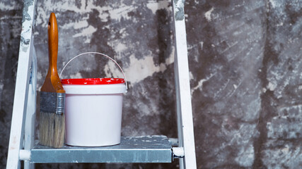 A paintbrush and a bucket of paint stand on a metal stepladder against an untreated concrete wall with remnants of white paint. Background