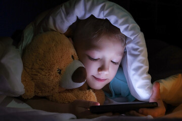 happy boy lies with toy bear in bed under a blanket and using a digital tablet smartphone device in the dark.